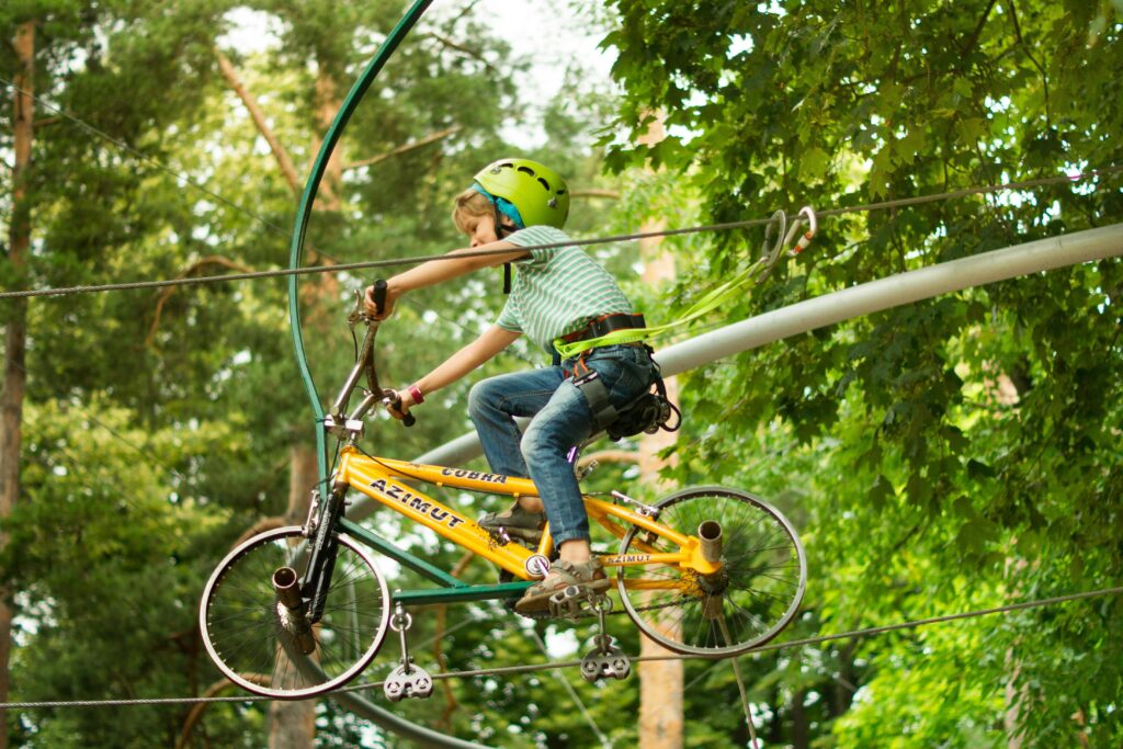 A child wearing a helmet rides a bike on a zipline in an adventure park surrounded by trees.